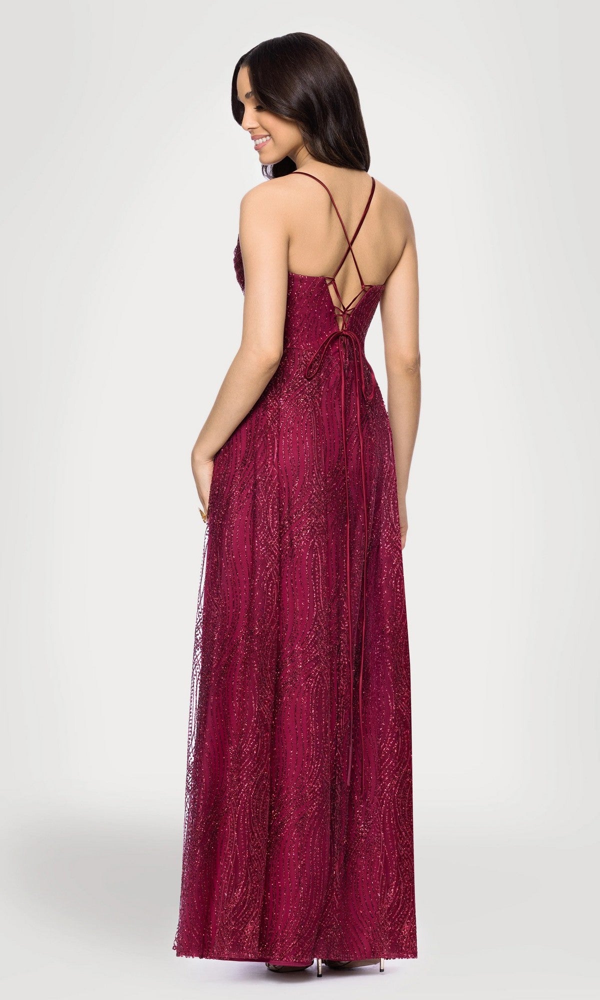 A woman stands smiling with her head turned to the side, wearing the Blondie Nites 5814BN long prom dress—a sleeveless, floor-length wine red glitter gown featuring crisscross straps and a lace-up back.